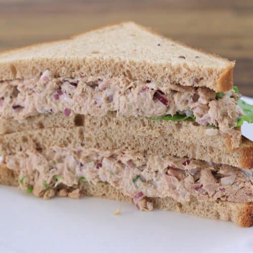 A sandwich made with two triangular slices of whole wheat bread, filled with a mixture of tuna, chopped red onions, and lettuce, is placed on a white plate. The sandwich is cut into halves and stacked on top of each other on a wooden table background.