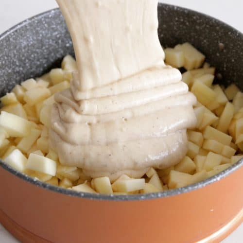 A thick, creamy mixture is being poured into a round baking pan filled with cubed potatoes. The pan has a two-tone color, with a non-stick gray interior and a copper-colored exterior. The background is a plain, light countertop.