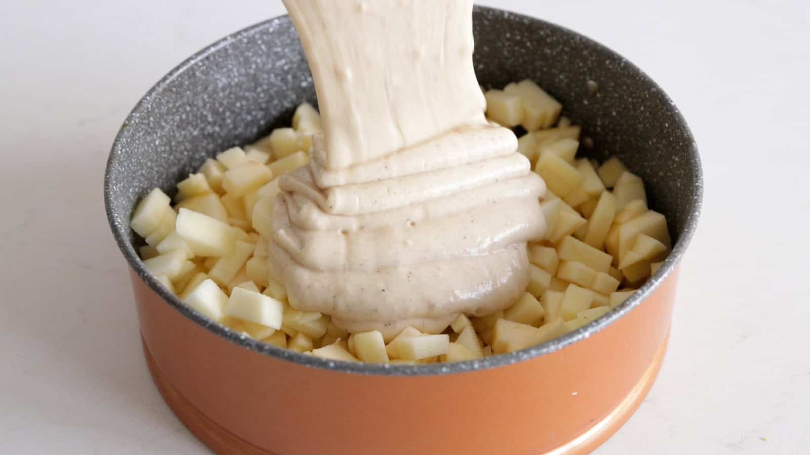 A thick, creamy mixture is being poured into a round baking pan filled with cubed potatoes. The pan has a two-tone color, with a non-stick gray interior and a copper-colored exterior. The background is a plain, light countertop.