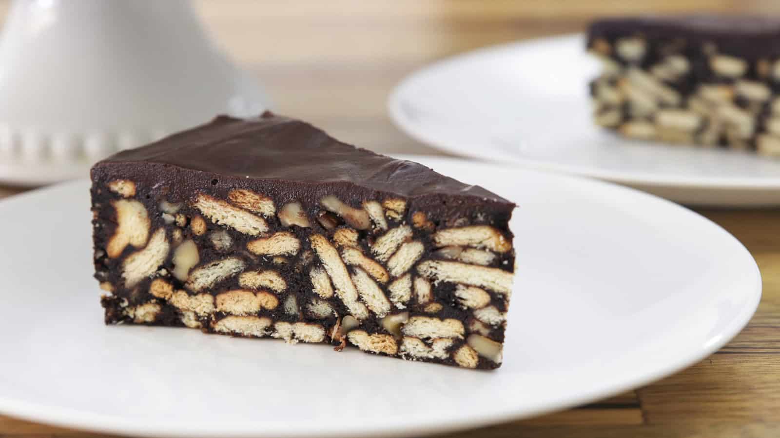 A slice of chocolate biscuit cake on a white plate. The cake has a rich, chocolatey exterior with visible pieces of biscuits embedded in it. Another slice of the same cake is blurred in the background on a similar plate.