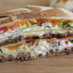 A close-up image of a cross-section of a burrito supreme. The layers reveal ground beef, melted cheese, diced tomatoes, shredded lettuce, sour cream, and a folded, crispy tortilla. The vibrant colors and textures of the fillings are clearly visible.