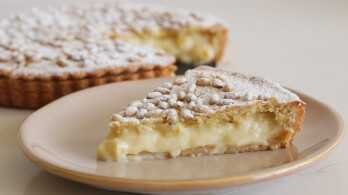 A close-up of a slice of lemon cream pie on a beige plate. The pie has a golden brown crust and is topped with powdered sugar. In the background, the whole pie, with a conspicuously missing slice, is partially visible.