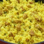 A close-up of a black frying pan filled with vibrant yellow rice mixed with chunks of chicken and green peas. The rice is evenly spread across the pan, with pieces of chicken distributed throughout. The wooden surface underneath the pan is partially visible.