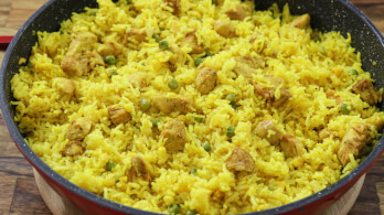 A close-up of a black frying pan filled with vibrant yellow rice mixed with chunks of chicken and green peas. The rice is evenly spread across the pan, with pieces of chicken distributed throughout. The wooden surface underneath the pan is partially visible.