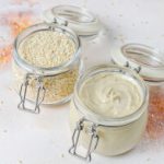 Two glass jars with metal clasps sit on a light surface. One jar is filled with sesame seeds, while the other contains creamy tahini. A few sesame seeds are scattered around the jars, adding to the overall rustic and natural presentation.