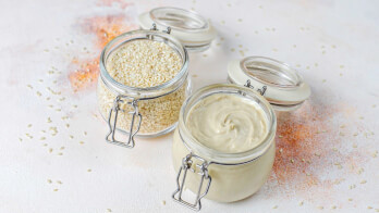 Two glass jars with metal clasps sit on a light surface. One jar is filled with sesame seeds, while the other contains creamy tahini. A few sesame seeds are scattered around the jars, adding to the overall rustic and natural presentation.