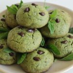 A plate of green cookies with chocolate chips arranged in a pile, garnished with fresh mint leaves. The cookies have a cracked, textured surface and look soft and freshly baked. The plate is set on a light, neutral background.