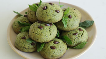 A plate of green cookies with chocolate chips arranged in a pile, garnished with fresh mint leaves. The cookies have a cracked, textured surface and look soft and freshly baked. The plate is set on a light, neutral background.