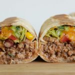 A close-up of a neatly cut burrito, showing its interior filled with ground meat, lettuce, beans, corn, and diced tomatoes. The burrito halves are placed on a wooden surface, displaying their vibrant and varied ingredients.