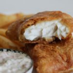 A close-up of golden brown battered fish fillets with a flaky white interior, served on a plate. In the background, there are crispy French fries, a wedge of lemon, and a side of creamy tartar sauce.