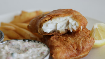 A close-up of golden brown battered fish fillets with a flaky white interior, served on a plate. In the background, there are crispy French fries, a wedge of lemon, and a side of creamy tartar sauce.