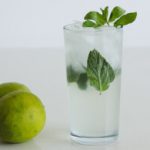 A tall glass filled with a clear drink, ice cubes, and garnished with mint leaves. Two whole limes are placed beside the glass on a white surface. The background is plain white.