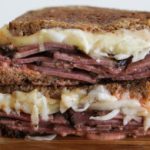 A close-up of a sandwich on a wooden surface. The sandwich is made with rye bread and filled with sliced corned beef, melted Swiss cheese, sauerkraut, and dressing, resembling a Reuben sandwich. The sandwich is cut in half, with both halves stacked.