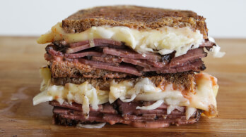 A close-up of a sandwich on a wooden surface. The sandwich is made with rye bread and filled with sliced corned beef, melted Swiss cheese, sauerkraut, and dressing, resembling a Reuben sandwich. The sandwich is cut in half, with both halves stacked.