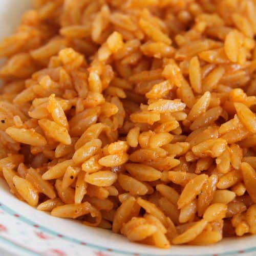 A close-up of a bowl filled with seasoned orzo pasta. The orzo is orange in color, indicating a sauce or seasoning has been mixed in. The edges of the bowl are decorated with a floral pattern. The dish appears warm and appetizing.