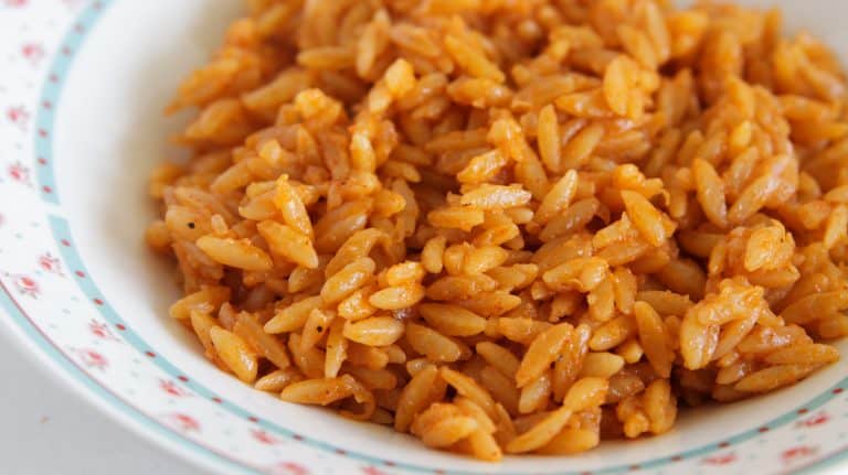 A close-up of a bowl filled with seasoned orzo pasta. The orzo is orange in color, indicating a sauce or seasoning has been mixed in. The edges of the bowl are decorated with a floral pattern. The dish appears warm and appetizing.