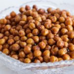A glass bowl filled with crispy roasted chickpeas, seasoned and browned, sits on a white surface. A few roasted chickpeas are scattered next to the bowl.