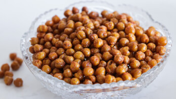 A glass bowl filled with crispy roasted chickpeas, seasoned and browned, sits on a white surface. A few roasted chickpeas are scattered next to the bowl.