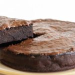 A rich, dense chocolate cake with one triangular slice being lifted out on a metal spatula. The cake has a glossy, chocolatey surface and appears moist and decadent, sitting on a light-colored plate.