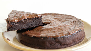 A rich, dense chocolate cake with one triangular slice being lifted out on a metal spatula. The cake has a glossy, chocolatey surface and appears moist and decadent, sitting on a light-colored plate.