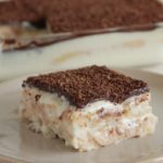 A slice of layered dessert featuring a creamy filling and a cocoa powder topping is served on a beige plate. The dessert dish with the remaining portion is visible in the background. The slice showcases distinct layers of filling and chocolate topping.