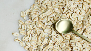 A close-up view of raw rolled oats spread out on a light surface with a small gold spoon resting on top.