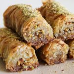 A plate of rolled baklava pastries, filled with chopped nuts and topped with a drizzle of honey and a sprinkling of ground pistachios. The layers of flaky, golden-brown phyllo dough are visible, showcasing a crunchy and syrupy texture.