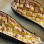 Two halves of roasted eggplant with a golden-brown, cross-hatch pattern on their flesh, placed on a baking sheet lined with parchment paper. The eggplant is cooked until soft and caramelized, with a slightly charred outer skin.