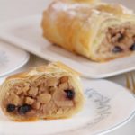 Close-up of sliced apple strudel on a white plate with gold-rimmed utensils and a striped cloth napkin beside it. In the background, a whole apple strudel rests on a rectangular white serving dish. The strudel's filling features apples and dark raisins.