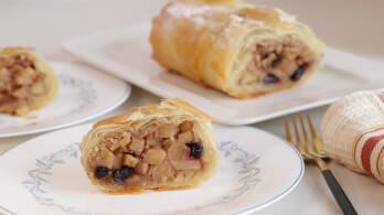 Close-up of sliced apple strudel on a white plate with gold-rimmed utensils and a striped cloth napkin beside it. In the background, a whole apple strudel rests on a rectangular white serving dish. The strudel's filling features apples and dark raisins.