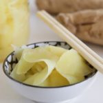 A small white bowl filled with pickled ginger slices is placed on a table. Next to the bowl are wooden chopsticks. In the background, there is a jar containing more pickled ginger and large pieces of ginger root.