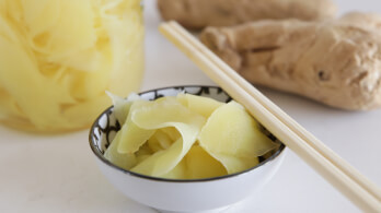 A small white bowl filled with pickled ginger slices is placed on a table. Next to the bowl are wooden chopsticks. In the background, there is a jar containing more pickled ginger and large pieces of ginger root.