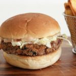 A vegan burger with a patty, shredded lettuce, and sauce in a bun on a wooden surface. To the right, there is a metal basket filled with golden French fries, with a few fries scattered on the surface nearby.