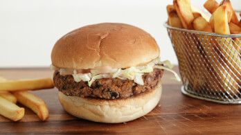 A vegan burger with a patty, shredded lettuce, and sauce in a bun on a wooden surface. To the right, there is a metal basket filled with golden French fries, with a few fries scattered on the surface nearby.