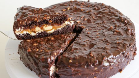 A chocolate cake with a glossy, textured chocolate exterior is shown with one slice being lifted out. The lifted slice reveals layers of chocolate cake, a creamy filling with visible nuts, and a caramel layer inside. The cake sits on a white surface.