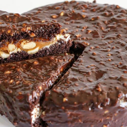 A chocolate cake with a glossy, textured chocolate exterior is shown with one slice being lifted out. The lifted slice reveals layers of chocolate cake, a creamy filling with visible nuts, and a caramel layer inside. The cake sits on a white surface.