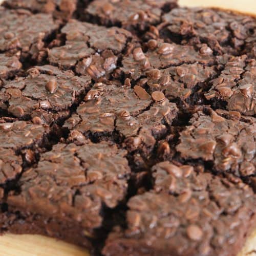 A tray of freshly baked brownies is cut into squares. The brownies appear rich and moist with a generous scattering of chocolate chips on top, giving them a textured, deliciously gooey look. They are placed on a wooden cutting board.