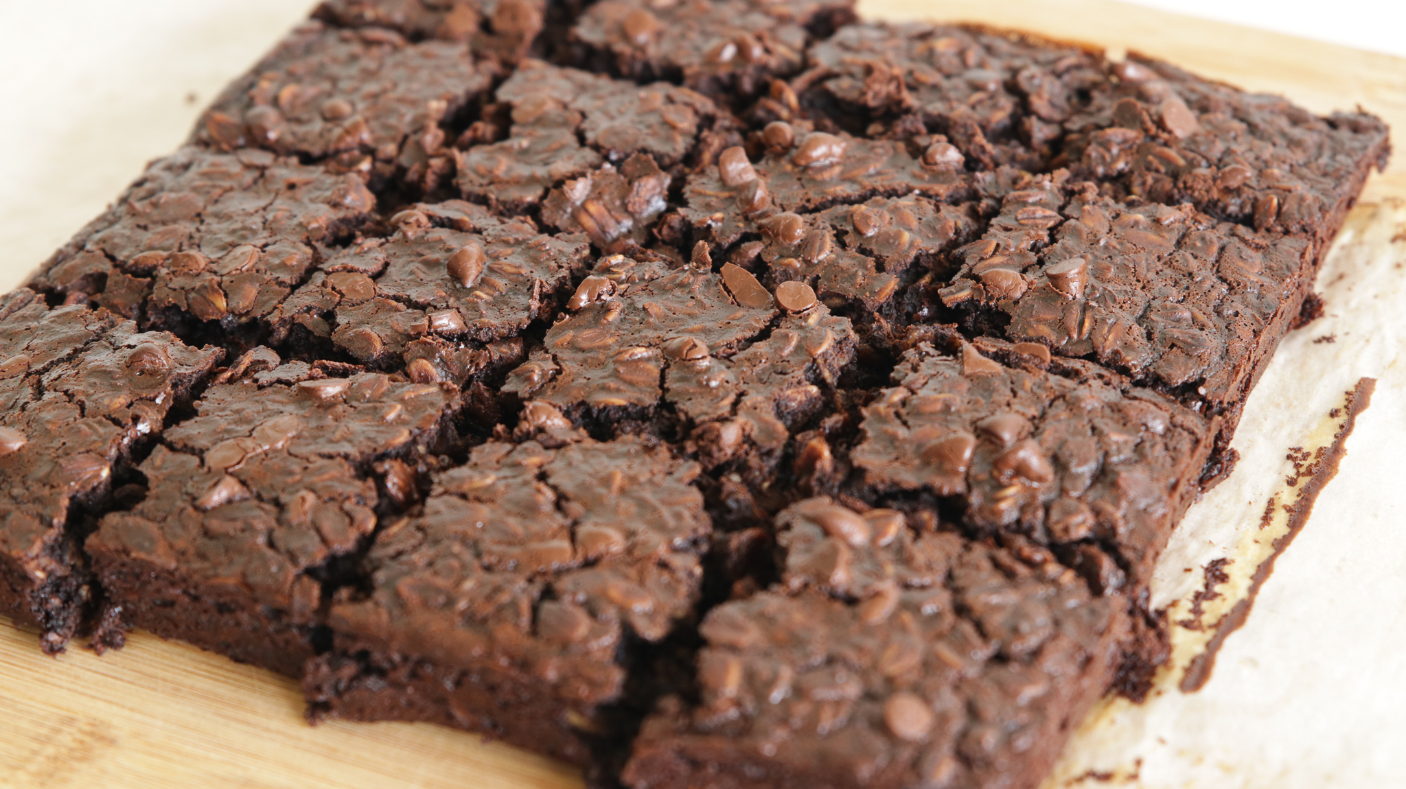 A tray of freshly baked brownies is cut into squares. The brownies appear rich and moist with a generous scattering of chocolate chips on top, giving them a textured, deliciously gooey look. They are placed on a wooden cutting board.