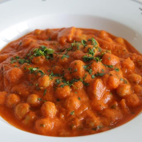 A white plate filled with a vibrant, red-orange curry made with chickpeas and spices, garnished with finely chopped fresh herbs. The dish appears rich and creamy, with a smooth, thick texture.