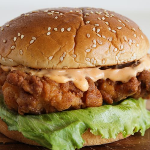 Close-up of a fried chicken sandwich on a sesame seed bun with a layer of crispy lettuce and a drizzle of creamy sauce, placed on a wooden surface. The sandwich showcases a golden-brown fried chicken fillet.