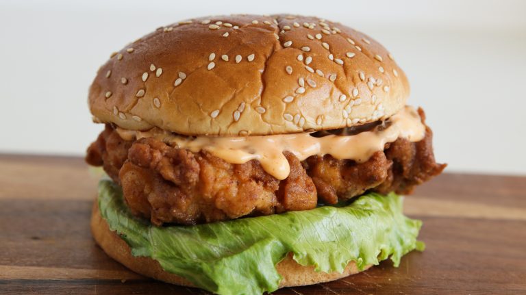 Close-up of a fried chicken sandwich on a sesame seed bun with a layer of crispy lettuce and a drizzle of creamy sauce, placed on a wooden surface. The sandwich showcases a golden-brown fried chicken fillet.