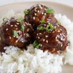 A beige plate holds a serving of white rice topped with three glazed meatballs. The meatballs are garnished with sesame seeds and chopped green onions, adding a touch of color and texture to the dish.