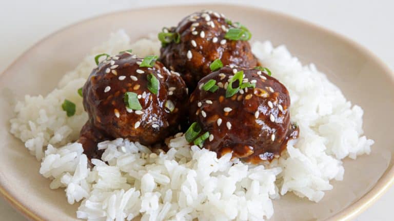 A beige plate holds a serving of white rice topped with three glazed meatballs. The meatballs are garnished with sesame seeds and chopped green onions, adding a touch of color and texture to the dish.