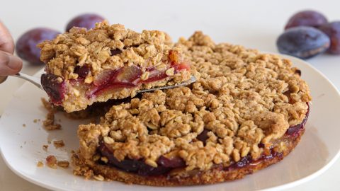 A hand lifts a slice of crumbly plum oat cake from a white plate. The cake has a golden, crunchy oat topping and a layer of cooked plums visible inside. Whole plums are blurred in the background.