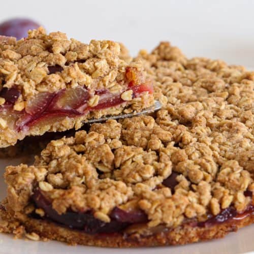 A hand lifts a slice of crumbly plum oat cake from a white plate. The cake has a golden, crunchy oat topping and a layer of cooked plums visible inside. Whole plums are blurred in the background.