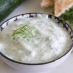 A bowl of creamy tzatziki sauce garnished with a small sprig of fresh dill sits on a light-colored surface. In the background, there is a cucumber, more fresh dill, and a piece of bread.