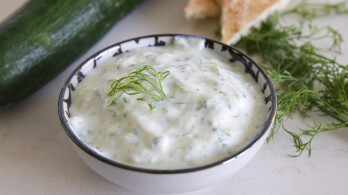 A bowl of creamy tzatziki sauce garnished with a small sprig of fresh dill sits on a light-colored surface. In the background, there is a cucumber, more fresh dill, and a piece of bread.