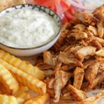 A plate of food featuring crinkle-cut fries, grilled seasoned chicken pieces, sliced tomatoes, onions, and a pita bread slice. A small bowl of creamy white dipping sauce is placed in the center.