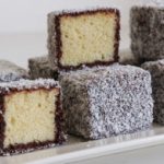 A plate of lamingtons, which are traditional Australian desserts. They are sponge cake squares coated in a layer of chocolate icing and rolled in desiccated coconut. Some of the lamingtons are cut open to reveal their soft, yellow sponge inside.