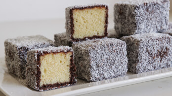 A plate of lamingtons, which are traditional Australian desserts. They are sponge cake squares coated in a layer of chocolate icing and rolled in desiccated coconut. Some of the lamingtons are cut open to reveal their soft, yellow sponge inside.